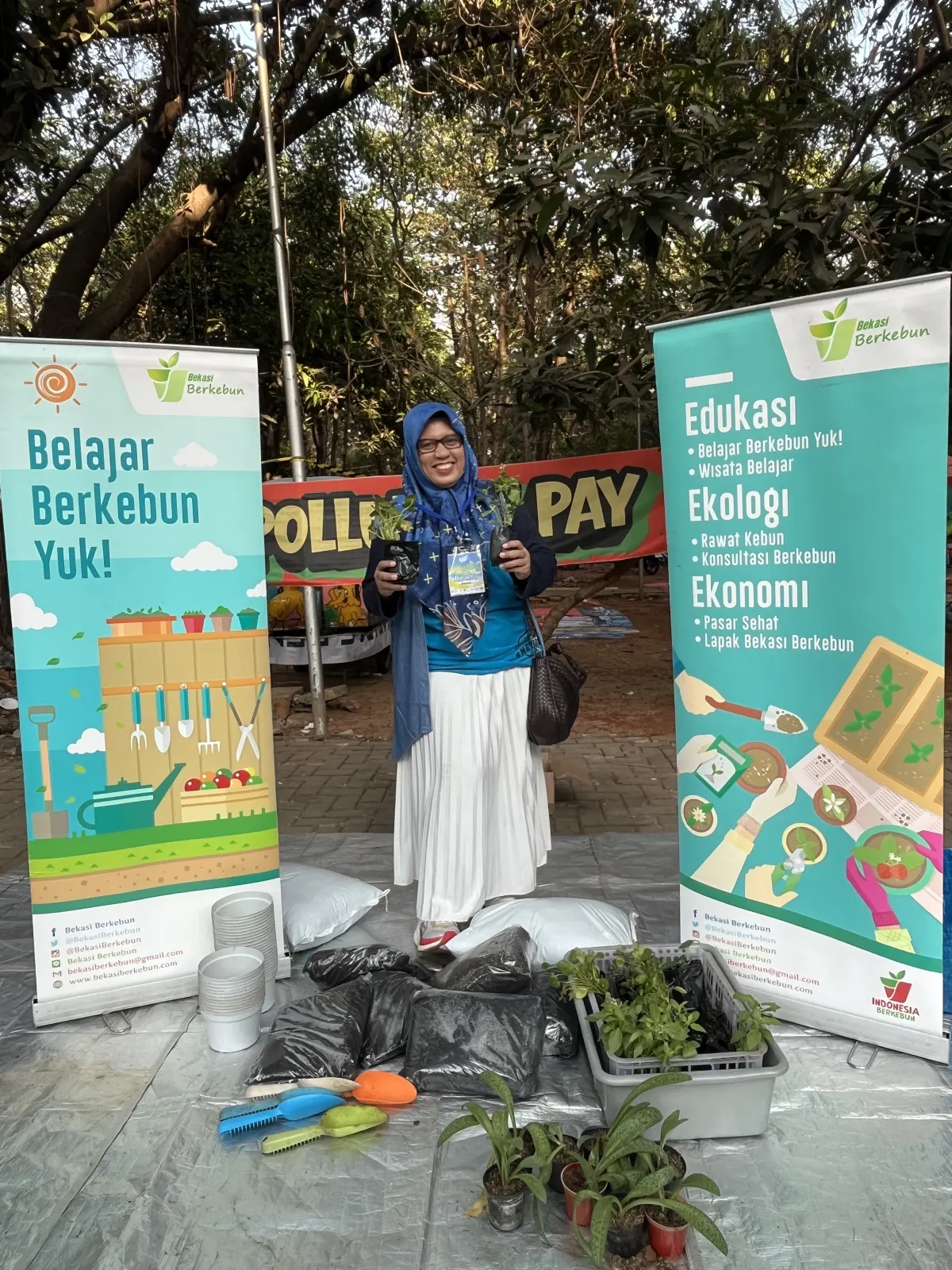 Seorang perempuan paruh baya sedang memegang dua bibit tanaman untuk dipindahkan ke pot sebagai kegiatan booth Bekasi Berkebun.