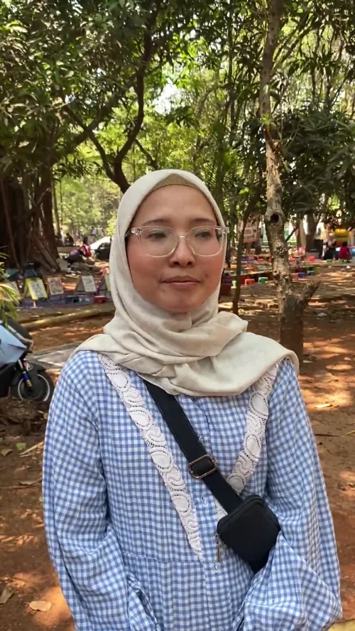 Erlyna dari Rangkul Bekasi berdiri di area berpohon Hutan Kota Bekasi, mengenakan hijab krem dan kemeja kotak-kotak biru putih.