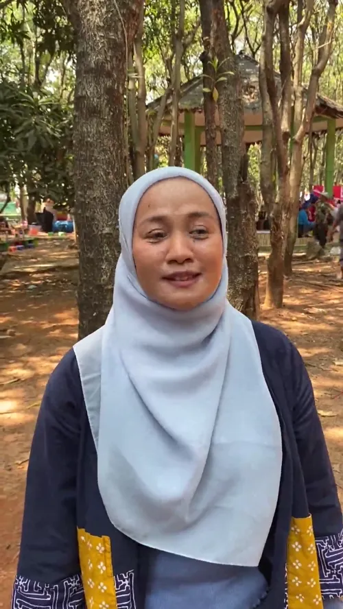 Rini dari Bekasi Read Aloud berdiri di area berpohon Hutan Kota Bekasi, mengenakan hijab abu-abu muda dan kardigan biru tua.