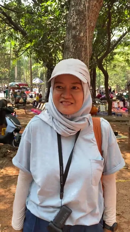 Winartania dari Bekasi Berkebun berdiri di area berpohon Hutan Kota Bekasi, mengenakan hijab putih bertopi dan kaus abu-abu lengan panjang.