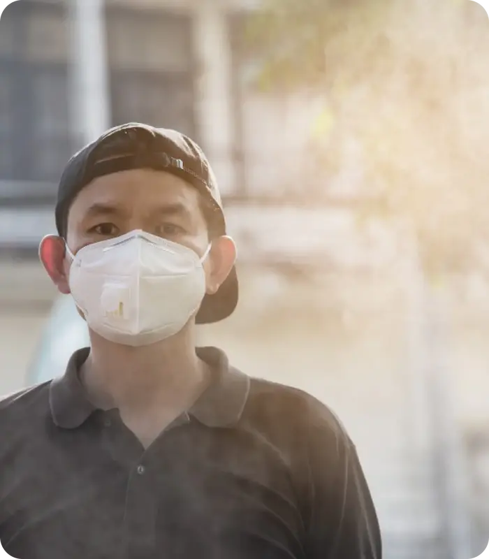 Seorang pria Asia mengenakan topi bisbol hitam terbalik dan masker wajah putih dengan memandang lurus ke depan dengan ekspresi serius. Ada sedikit kabut atau asap di udara di sekelilingnya, dan cahaya terang terlihat di sisi kanan gambar.