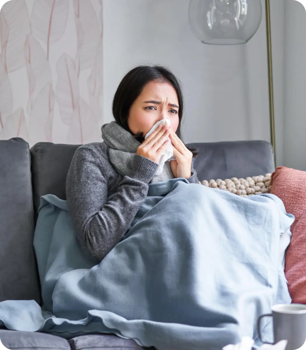 Seorang wanita Asia yang tampak tidak sehat, terbungkus selimut dan syal abu-abu, sedang duduk di sofa abu-abu dan menyeka hidungnya dengan tisu. Di sebelahnya ada bantal merah muda dengan rumbai. Latar belakangnya adalah dinding bermotif.