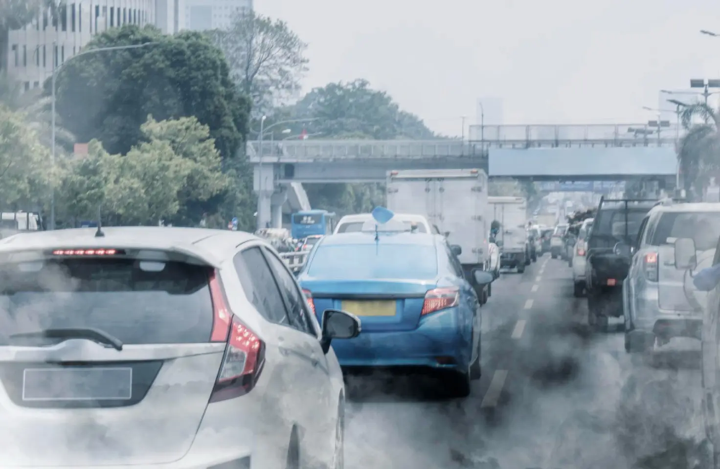 Pemandangan kabur dari kemacetan lalu lintas kota di jalan raya yang tertutup kabut atau asap tebal dari emisi knalpot, dengan banyak mobil, truk, dan jembatan layang di latar belakang.